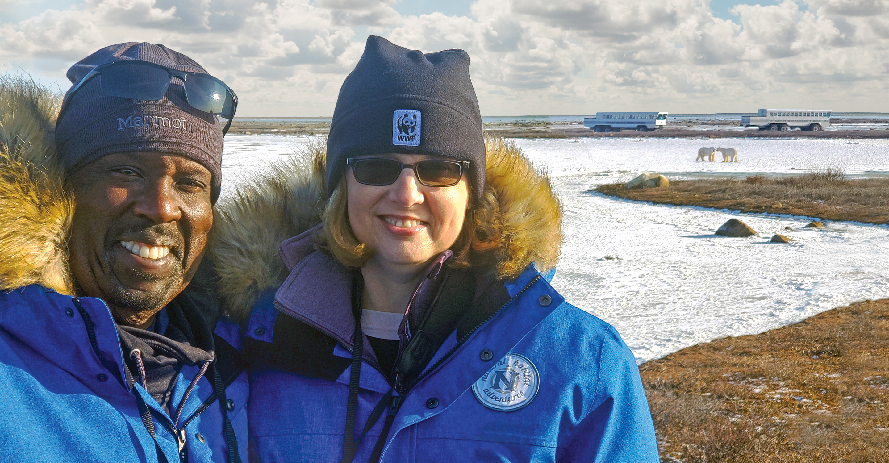 Two Natural Habitat Adventures travelers smile in front of two Polar Rovers and two polar bears on the tundra in Churchill, Manitoba, Canada