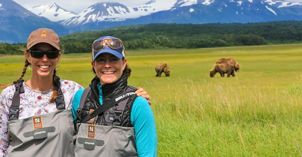 Two smiling travelers stand in front of three grazing brown bears in Katmai National Park, Alaska, USA