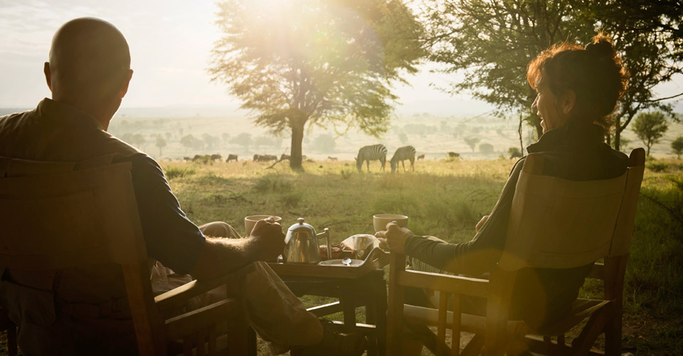 Travelers at Natural Habitat's Migration Base Camp sit on their porch with tea service and view zebra grazing, Serengeti National Park, Tanzania