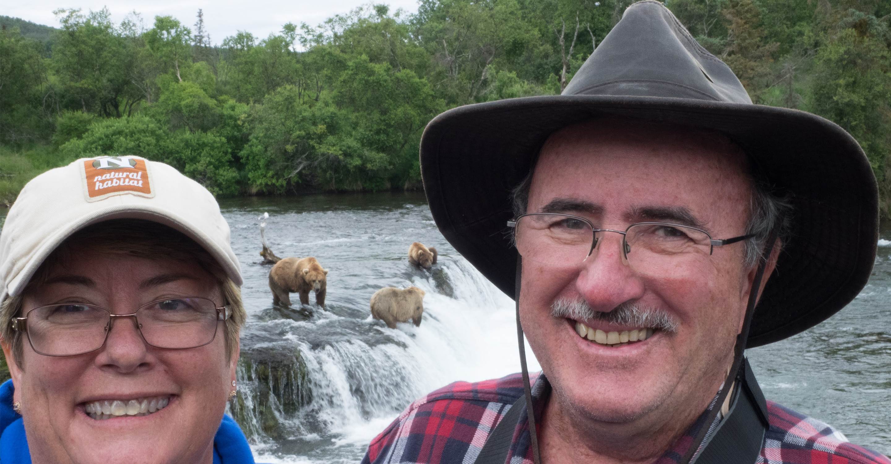 Travelers smile in front of brown bears standing in Brooks Falls, Alaska, USA